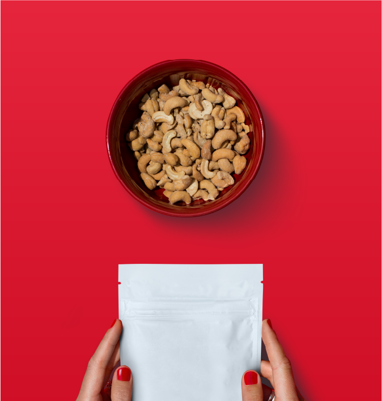 A bowl of cashew nuts on a red background with hands holding a white resealable pouch beneath it.